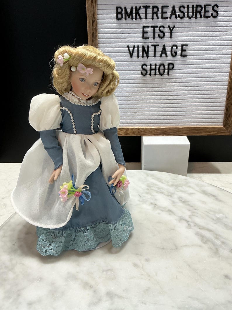 May include: A vintage porcelain doll with blonde hair and a blue dress with white accents. The doll has a floral headpiece and is holding a bouquet of flowers. The background includes a sign that reads "BMKTREASURES ETSY VINTAGE SHOP."