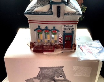 Op de afbeelding: Een keramische miniatuur van een barbershop met een met sneeuw bedekt dak en de tekst "Beard Barber". Het gebouw heeft een rode deur, een barbierpaal en decoratieve kransen. Het staat op een witte doos met een afgedrukte afbeelding van de winkel.
