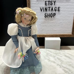 May include: A vintage porcelain doll with blonde hair and a blue and white dress. The doll has a floral hair accessory and is holding a bouquet of flowers. The text "BMKTREASURES ETSY VINTAGE SHOP" is in the background.