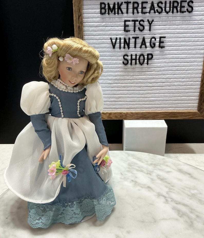 May include: A vintage porcelain doll with blonde hair and a blue and white dress. The doll has a floral hair accessory and is holding a bouquet of flowers. The text "BMKTREASURES ETSY VINTAGE SHOP" is in the background.