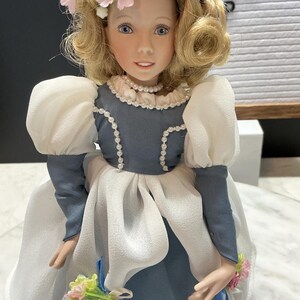 May include: A porcelain doll with blonde hair styled in curls and adorned with pink flowers. The doll wears a blue and white dress with a pearl necklace and holds a small bouquet of flowers. The doll's face has blue eyes and a gentle expression.