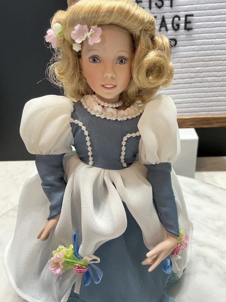 May include: A porcelain doll with blonde hair styled in curls and adorned with pink flowers. The doll wears a blue and white dress with a pearl necklace and holds a small bouquet of flowers. The doll's face has blue eyes and a gentle expression.