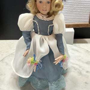 May include: A porcelain doll with blonde curly hair and blue eyes, wearing a blue and white dress with lace trim. The doll has a pearl necklace and holds small bouquets. The doll is a vintage item.