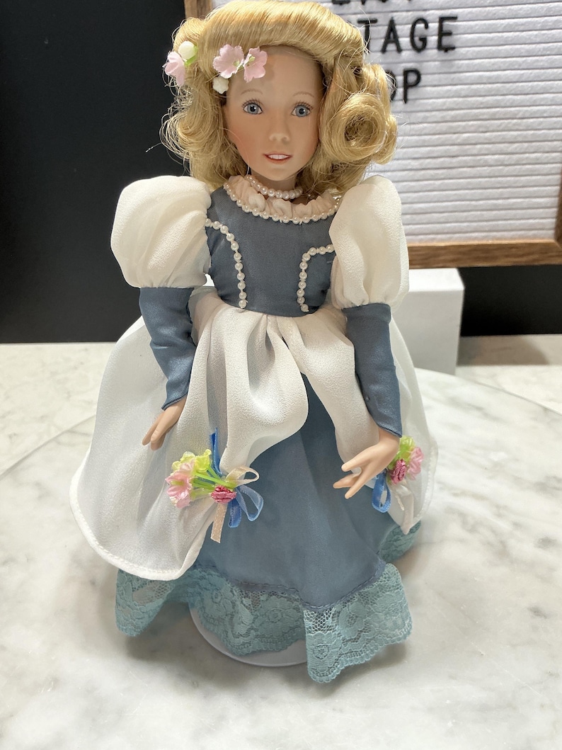 May include: A porcelain doll with blonde curly hair and blue eyes, wearing a blue and white dress with lace trim. The doll has a pearl necklace and holds small bouquets. The doll is a vintage item.