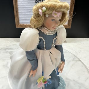 May include: A porcelain doll with blonde hair adorned with pink flowers. The doll wears a blue dress with white accents and a pearl necklace. The doll is holding a small bouquet of flowers. The background includes a sign that says "VINTAGE SHOP".