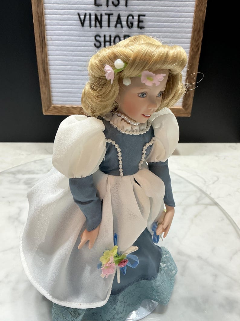 May include: A porcelain doll with blonde hair adorned with pink flowers. The doll wears a blue dress with white accents and a pearl necklace. The doll is holding a small bouquet of flowers. The background includes a sign that says "VINTAGE SHOP".