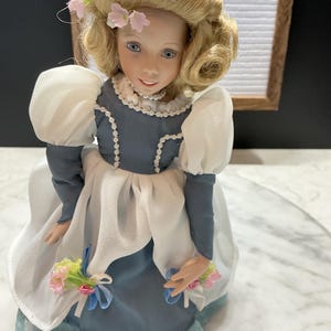 May include: A porcelain doll with blonde hair and blue eyes, wearing a blue and white dress with lace trim. The doll has a pearl necklace and holds a bouquet of flowers. The word "ETSY" is visible in the background.