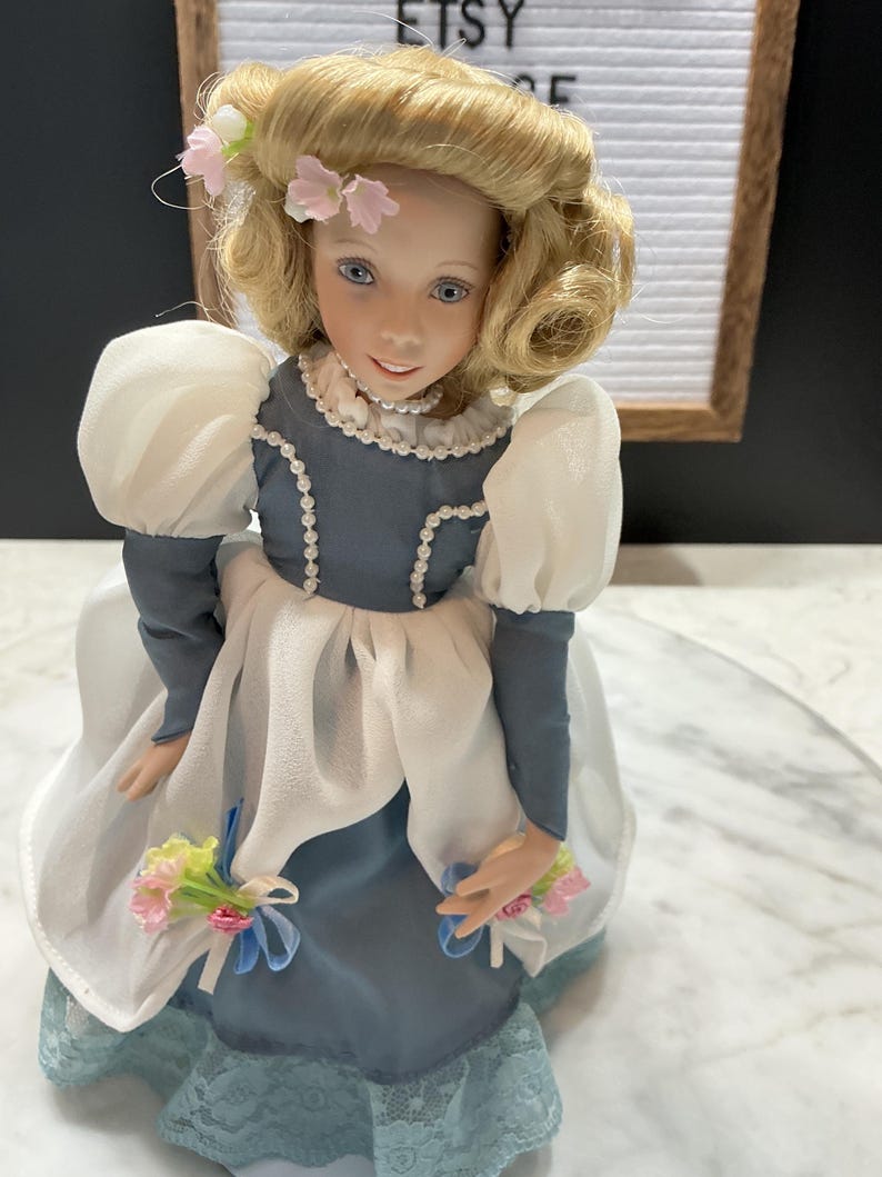 May include: A porcelain doll with blonde hair and blue eyes, wearing a blue and white dress with lace trim. The doll has a pearl necklace and holds a bouquet of flowers. The word "ETSY" is visible in the background.