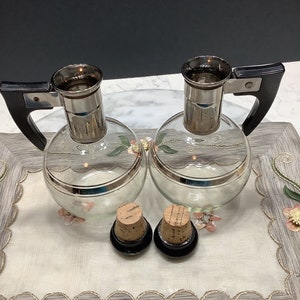 May include: Two clear glass decanters with black handles and silver tops. The decanters have a silver band around the middle. Two cork stoppers are in the foreground.