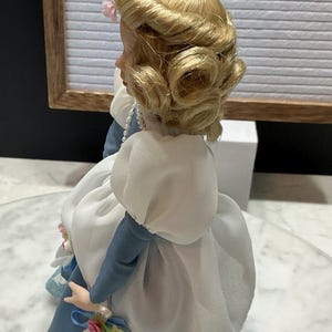May include: A vintage porcelain doll with blonde hair styled in curls, wearing a blue and white dress. The doll has a pearl necklace and is holding a small bouquet. The background includes a sign that says "ETSY VINTAGE SHOP."