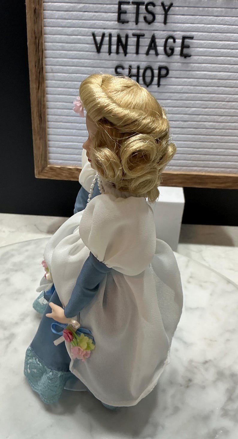 May include: A vintage porcelain doll with blonde hair styled in curls, wearing a blue and white dress. The doll has a pearl necklace and is holding a small bouquet. The background includes a sign that says "ETSY VINTAGE SHOP."