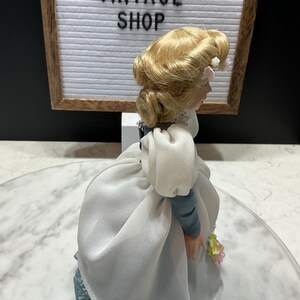 May include: A vintage porcelain doll with blonde hair styled in an updo, wearing a white dress with blue sleeves and a blue lace skirt. The doll is holding a small bouquet of flowers. The background includes a sign that reads "ETSY VINTAGE SHOP."