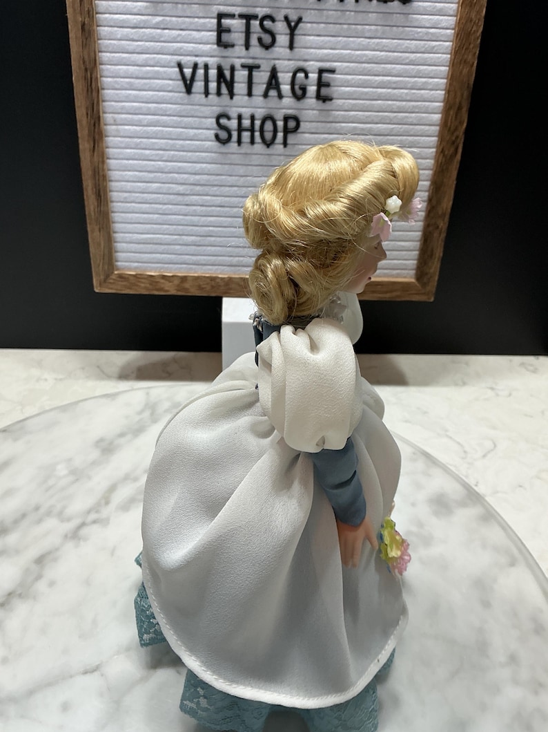 May include: A vintage porcelain doll with blonde hair styled in an updo, wearing a white dress with blue sleeves and a blue lace skirt. The doll is holding a small bouquet of flowers. The background includes a sign that reads "ETSY VINTAGE SHOP."