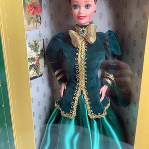 May include: A doll in a box, wearing a long, emerald green dress with a gold-trimmed bodice and a gold bow. The doll has brown hair and is in a decorative box with Christmas-themed artwork.