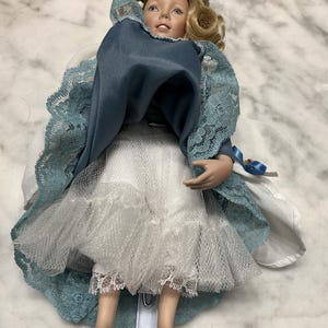 May include: A porcelain doll with blonde hair and blue eyes, wearing a blue dress with lace sleeves and a white skirt. The doll has a delicate face and is standing on a clear plastic stand.