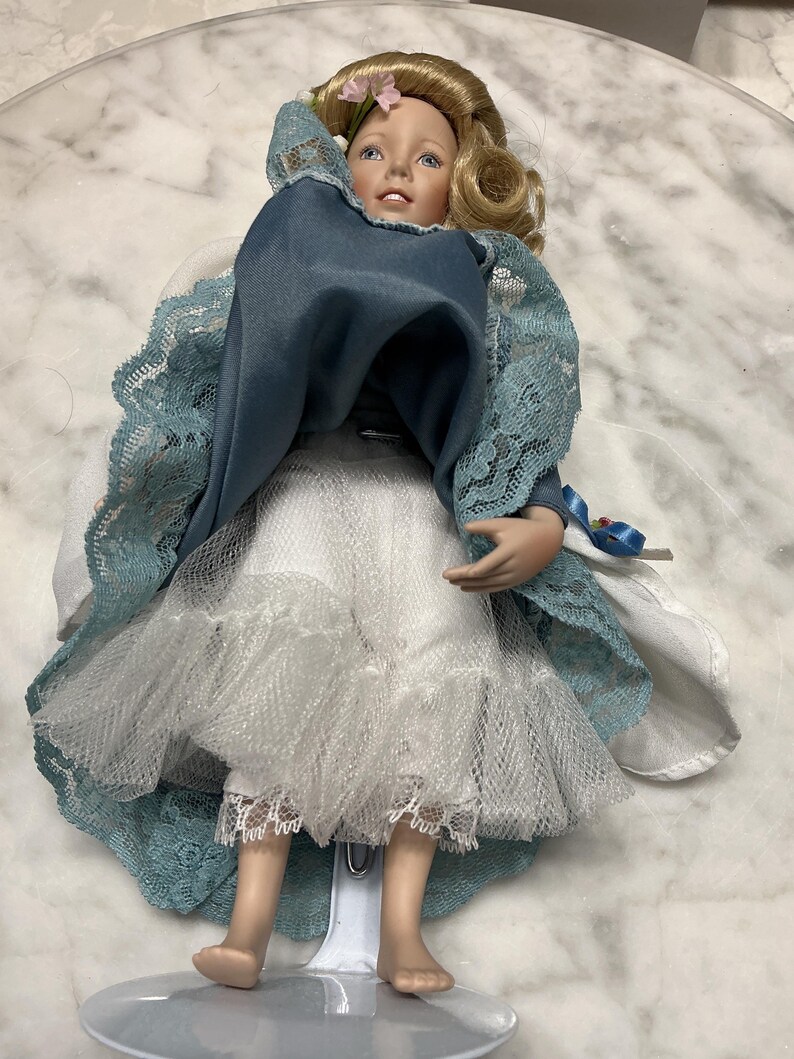 May include: A porcelain doll with blonde hair and blue eyes, wearing a blue dress with lace sleeves and a white skirt. The doll has a delicate face and is standing on a clear plastic stand.
