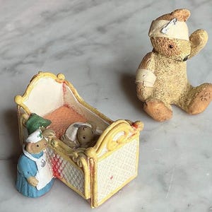 May include: Figurines of teddy bears in a medical theme. One bear is sitting with a bandage on its head and arm. Another bear is in a crib with a nurse bear tending to it. The crib is yellow and white.
