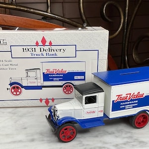 May include: A die-cast metal 1931 Delivery Truck Bank, 1/34 scale, with rubber tires. The white truck has blue and black accents, red wheels, and the True Value Hardware logo. The box is also visible.