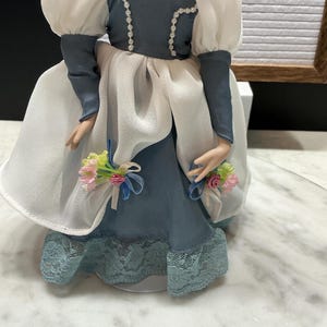 May include: A porcelain doll in a blue and white dress with a lace hem. The doll has a pearl necklace and a blue bodice with white trim. The doll's sleeves are white and puffy. Small bouquets of pink and yellow flowers are attached to the doll's dress.