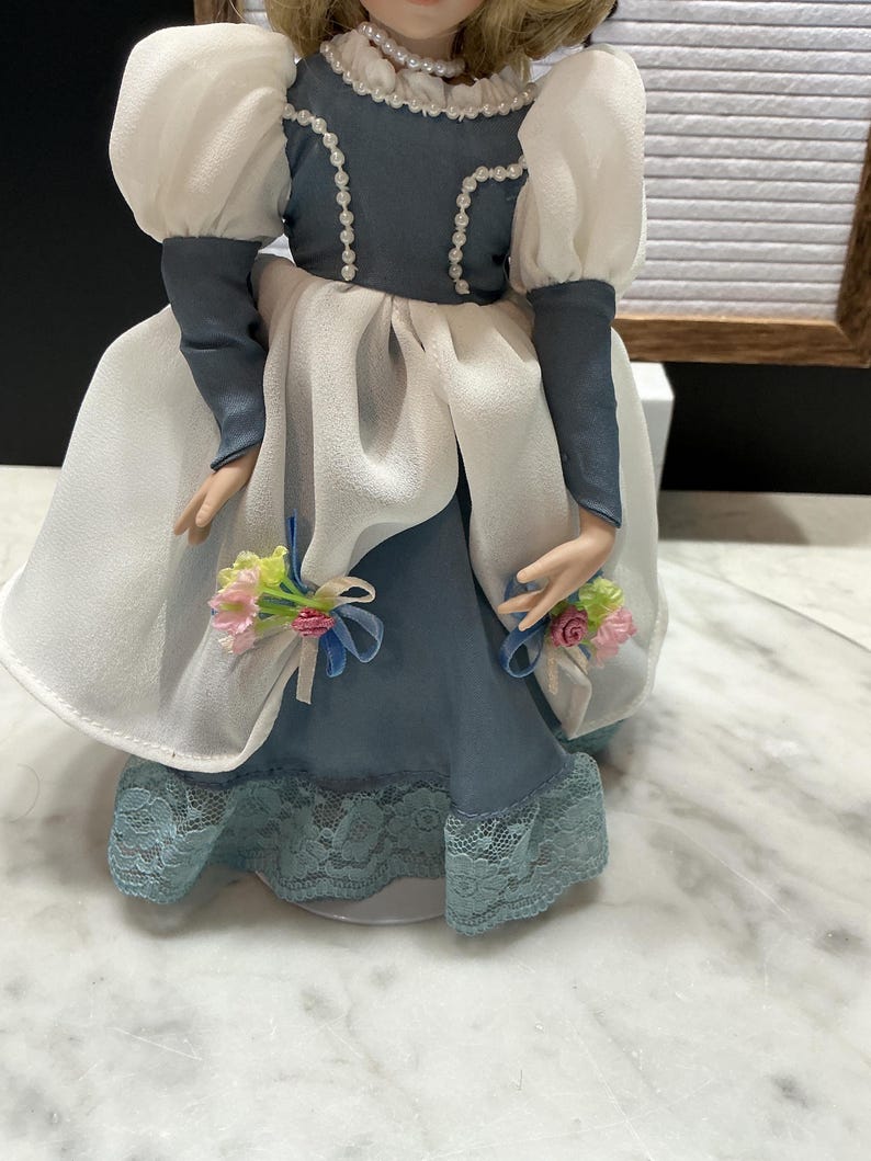 May include: A porcelain doll in a blue and white dress with a lace hem. The doll has a pearl necklace and a blue bodice with white trim. The doll's sleeves are white and puffy. Small bouquets of pink and yellow flowers are attached to the doll's dress.