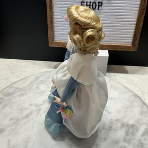 May include: A vintage porcelain doll with blonde hair styled in curls, wearing a blue and white dress. The doll is holding a small bouquet of flowers. The background includes a sign that reads "ETSY VINTAGE SHOP".