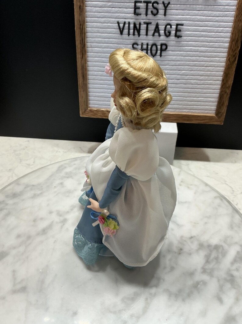 May include: A vintage porcelain doll with blonde hair styled in curls, wearing a blue and white dress. The doll is holding a small bouquet of flowers. The background includes a sign that reads "ETSY VINTAGE SHOP".