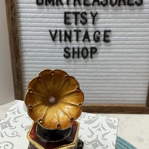 May include: A miniature gramophone figurine featuring a gold-toned horn with small embedded crystals, set atop a hexagonal base with deep red and green enamel detailing. The background displays a letter board with the text BMKTREASURES ETSY VINTAGE SHOP.