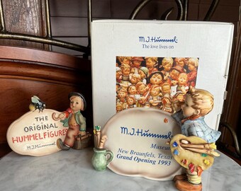 Może przedstawiać: Ekspozycja figurek Hummel i pudełka. Jedna figurka trzyma pędzel i paletę. Kolejna figurka znajduje się obok znaku z napisem "The Original Hummel Figures". Na pudełku znajduje się napis "M.J. Hummel" i "The love lives on."