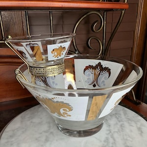 May include: A vintage glass punch bowl set with a smaller bowl resting inside. The bowls feature gold and white geometric designs with ornate floral accents. The set includes a gold-toned metal stand and handle.