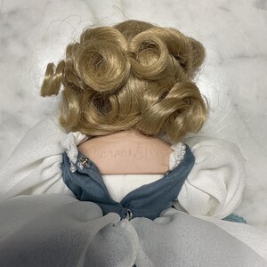 May include: A vintage doll with blonde, curly hair. The doll is wearing a blue and white dress with a pearl-like trim. The doll's name is printed on the back of the neck.