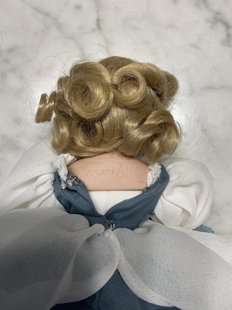 May include: A vintage doll with blonde, curly hair. The doll is wearing a blue and white dress with a pearl-like trim. The doll's name is printed on the back of the neck.