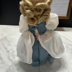 May include: A vintage doll with blonde, curled hair and a blue dress with white sleeves. The doll is standing on a clear surface. The doll's dress has a bow and decorative buttons.