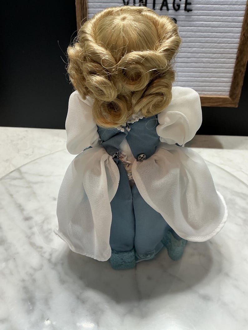 May include: A vintage doll with blonde, curled hair and a blue dress with white sleeves. The doll is standing on a clear surface. The doll's dress has a bow and decorative buttons.