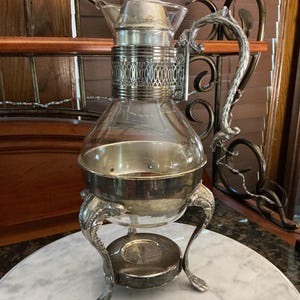 May include: A clear glass carafe with a silver-plated metal frame, featuring an ornate handle, a decorative filigree collar, a matching lid with a finial, and three curved legs that support the base of the glass vessel.