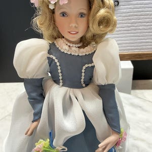 May include: A porcelain doll with blonde hair styled in curls and adorned with pink flowers. The doll wears a blue and white dress with a pearl necklace and holds a small bouquet. The doll's face has blue eyes and a gentle expression.