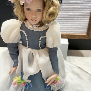 May include: A vintage porcelain doll with blonde hair, blue eyes, and a delicate floral crown. The doll wears a blue and white dress with puff sleeves and holds small bouquets. The word "AGE" is visible in the background.