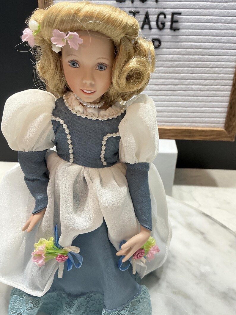 May include: A vintage porcelain doll with blonde hair, blue eyes, and a delicate floral crown. The doll wears a blue and white dress with puff sleeves and holds small bouquets. The word "AGE" is visible in the background.
