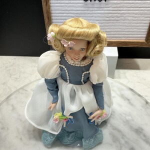 May include: A porcelain doll with blonde hair and a blue dress. The doll has a delicate face with blue eyes and is adorned with pink flowers. The dress features white sleeves and a lace trim. The doll is holding a small bouquet of flowers.