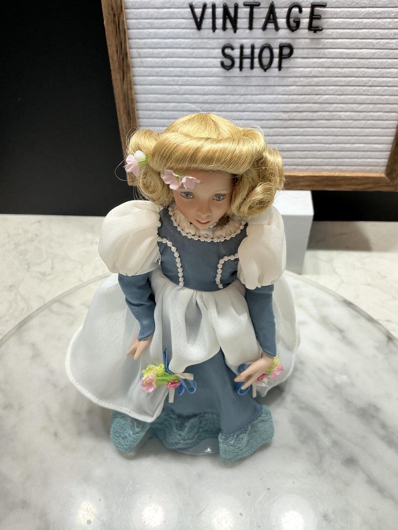 May include: A porcelain doll with blonde hair and a blue dress. The doll has a delicate face with blue eyes and is adorned with pink flowers. The dress features white sleeves and a lace trim. The doll is holding a small bouquet of flowers.