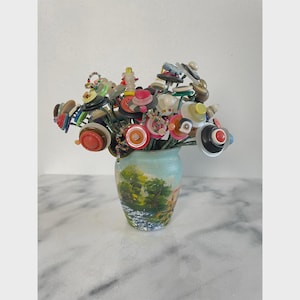 May include: A bouquet of button flowers in a painted ceramic vase. The button flowers are made of various colors and sizes of buttons. The vase is light blue with a landscape painting. The arrangement is on a white marble surface.