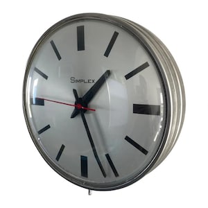 May include: A round, silver-toned Simplex clock with a white face. Black rectangular hour markers and black hands indicate the time, with a red second hand. The clock has a glass cover and a ridged metal frame.