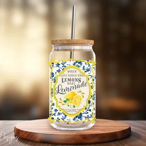 May include: A clear glass drinking cup with a wooden lid and a metal straw. The cup features a label with the text "When life gives you lemons make lemonade" and a lemon graphic. The design includes blue floral accents.