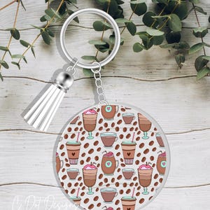 May include: A round, clear keychain with a white tassel. The keychain features a pattern of coffee cups, coffee beans, and hearts on a light brown background.