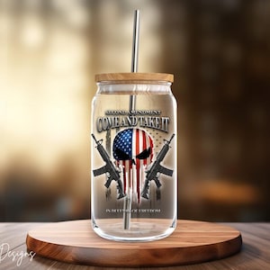 May include: A clear glass mason jar with a wooden lid and a straw. The jar has a graphic of a skull with crossed rifles and an American flag. The text "Second Amendment Come and Take It" is printed on the jar. The text "In Defense of Freedom" is printed below the graphic.