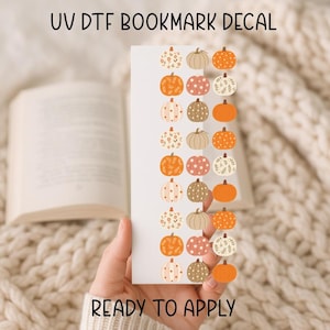 May include: A white bookmark decal with a repeating pattern of colorful pumpkins. The pumpkins are in various designs and colors, including orange, beige, and pink. Text at the top reads "UV DTF BOOKMARK DECAL" and at the bottom "READY TO APPLY".