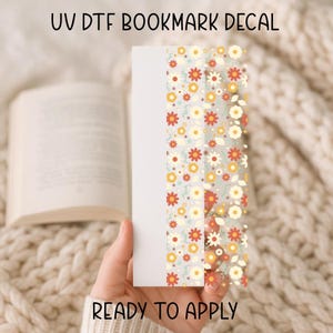 May include: A white bookmark with a floral design featuring red, yellow, and white flowers. The text "UV DTF BOOKMARK DECAL" and "READY TO APPLY" are visible. The bookmark is held in a person's hand.