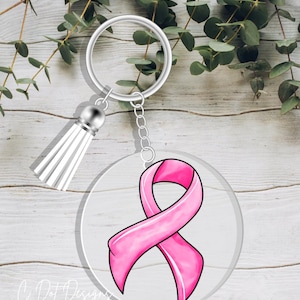 May include: A clear, round keychain with a pink ribbon design, silver hardware, and a white tassel. The ribbon is a symbol of breast cancer awareness. The keychain is a thoughtful gift or accessory.