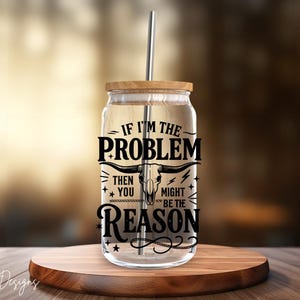May include: A clear glass tumbler with a wooden lid and a stainless steel straw. The tumbler features black text that reads "IF I'M THE PROBLEM THEN YOU MIGHT BE THE REASON" with a bull skull graphic. The tumbler sits on a wooden surface.