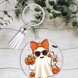 May include: A Halloween-themed keychain featuring a cartoon ghost with sunglasses, an orange bow, and a purse. The ghost is holding a pumpkin and a coffee cup. The keychain has a silver ring and a white tassel. The design includes pumpkins and a bat.