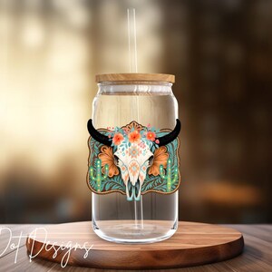 May include: A clear glass tumbler with a wooden lid and straw. The tumbler features a decorative design of a bull skull with floral accents and cactus on a turquoise background. The tumbler sits on a wooden coaster.